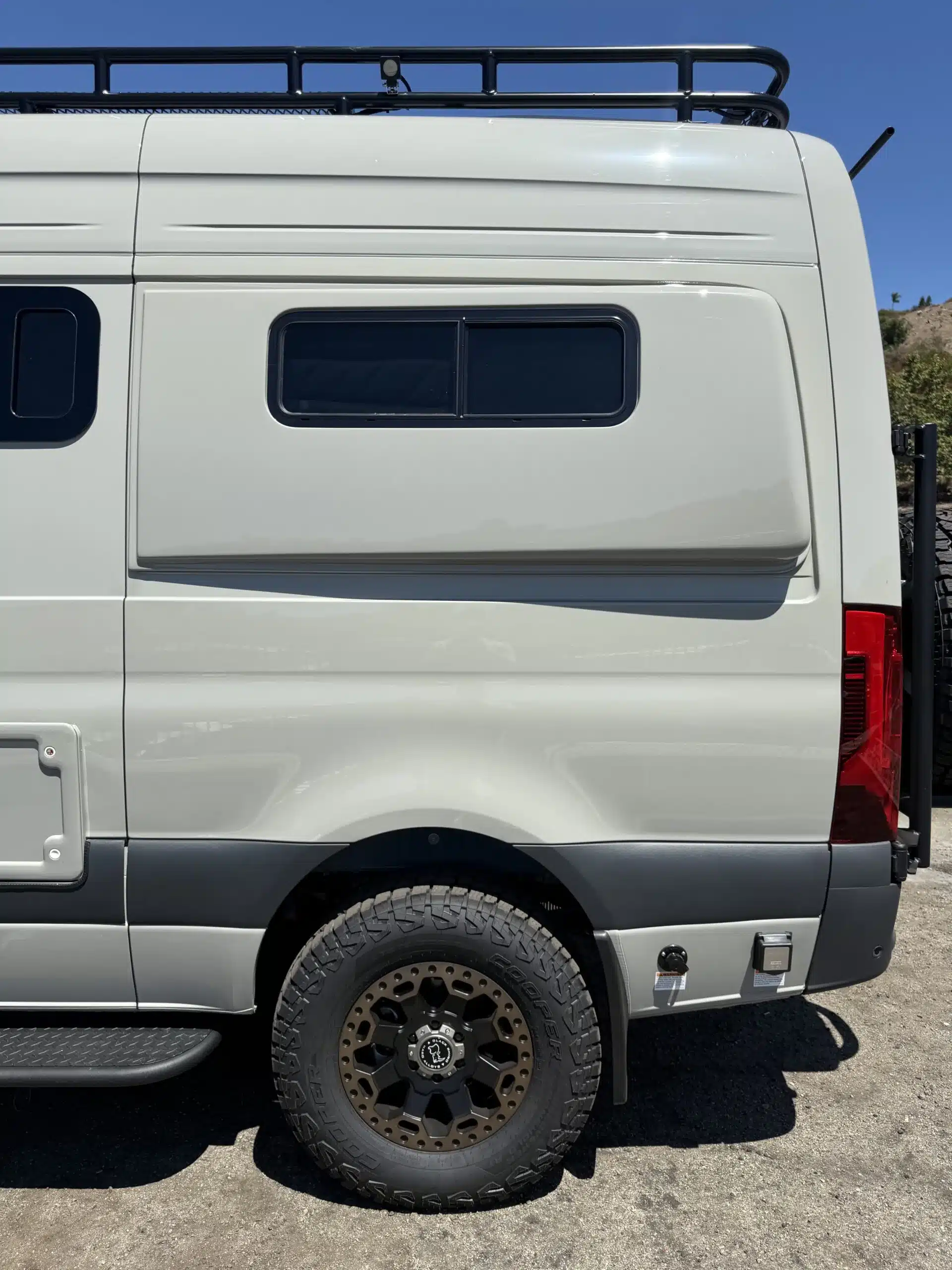 Detailansicht eines weißen Kastenwagens mit robusten Offroad-Reifen und Dachgepäckträger bei Sonnenschein