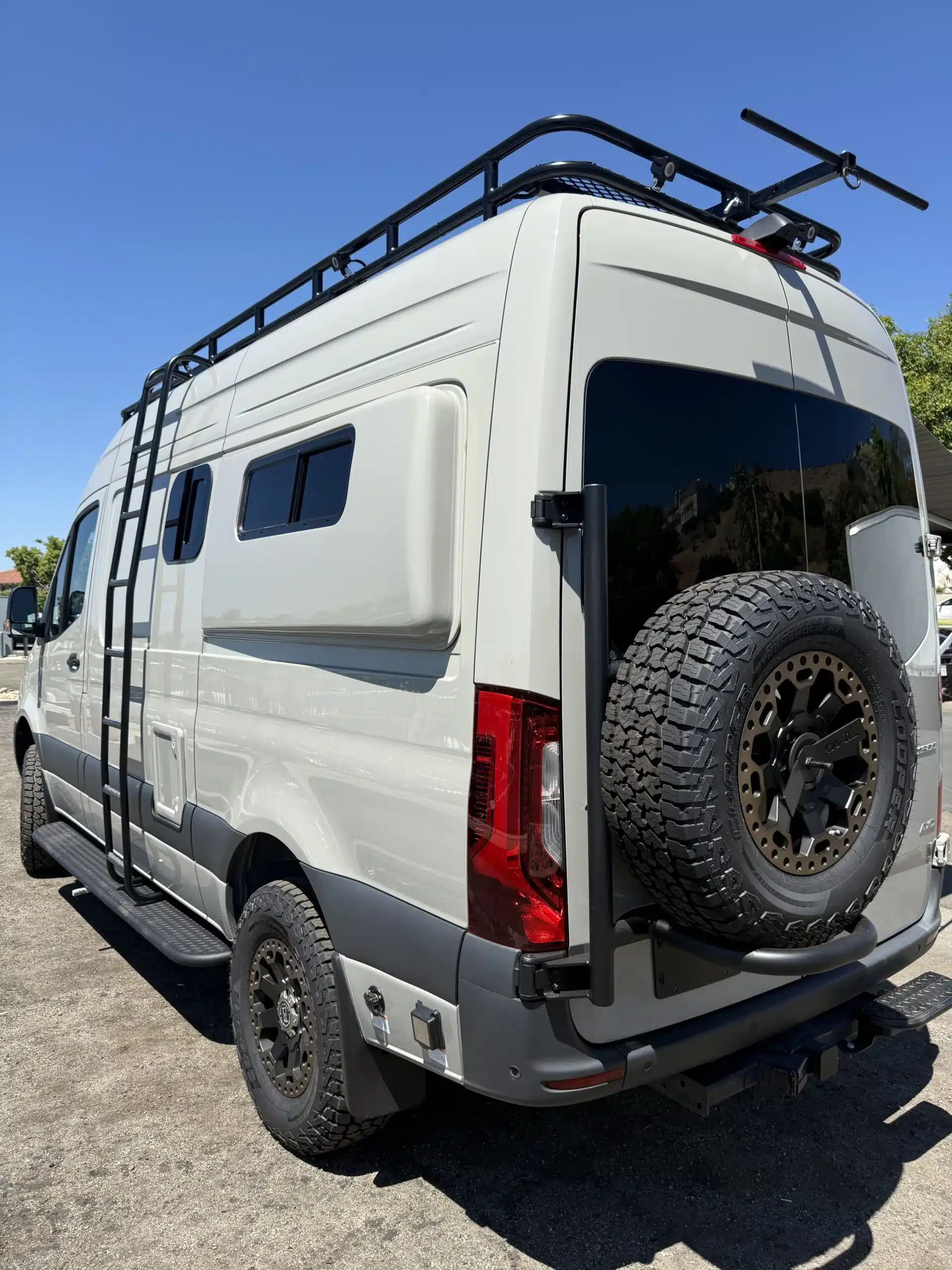 Der Trail-Hob Adventure-Van auf Basis des Sprinter 144 AWD, ausgestattet mit Dachträger, Heckleiter und großem Ersatzrad, von hinten fotografiert.