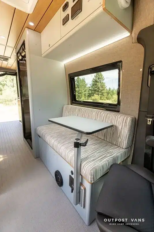 Sitzbank mit Klapptisch im Innenraum eines modernen Offroad-Campervans, Fensterblick ins Grüne, hochwertige Ausbaumaterialien.