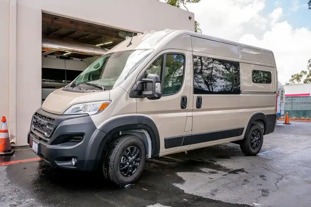 Beiger Avalon Campervan von Vamonos Vans vor einer Werkstatt, seitliche Ansicht.
