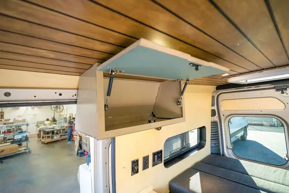 Aufgeklappter Hängeschrank im Innenraum eines Avalon Kastenwagen-Campers mit Holzdecke und Sicht auf modulare Bettenlösung.