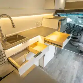 Blick auf den Küchenbereich mit Spüle, Auszügen und Kühlschrank im Innenraum des Turq Campervans von Vamonos Vans
