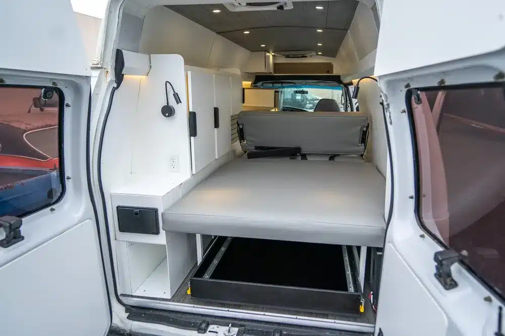Blick in den Innenraum eines Turq Campervans mit ausklappbarem Bett und weißen Aluminium-Schrankmodulen.