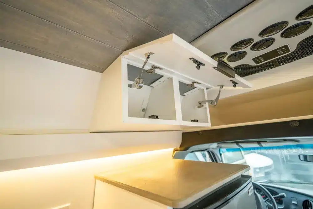 Geöffneter Hängeschrank mit Aluminium-Bauweise und Übersicht der Innenausstattung im Vamonos Vans Turq Campervan