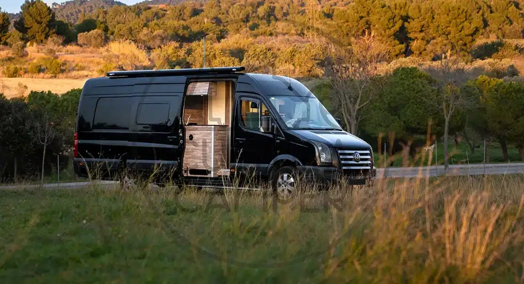 Schwarzer Volkswagen Crafter L4H2 Campervan mit Seitentür, abgestellt in einer grünen Landschaft bei Sonnenuntergang.