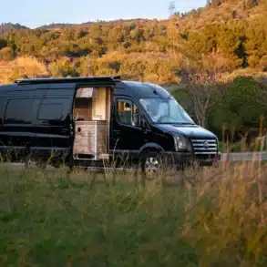 Schwarzer Volkswagen Crafter L4H2 Campervan mit Seitentür, abgestellt in einer grünen Landschaft bei Sonnenuntergang.