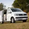 VW Caravelle T6 Camper auf einer Wiese mit geöffneter Schiebetür, Campingausbau im Innenraum sichtbar.