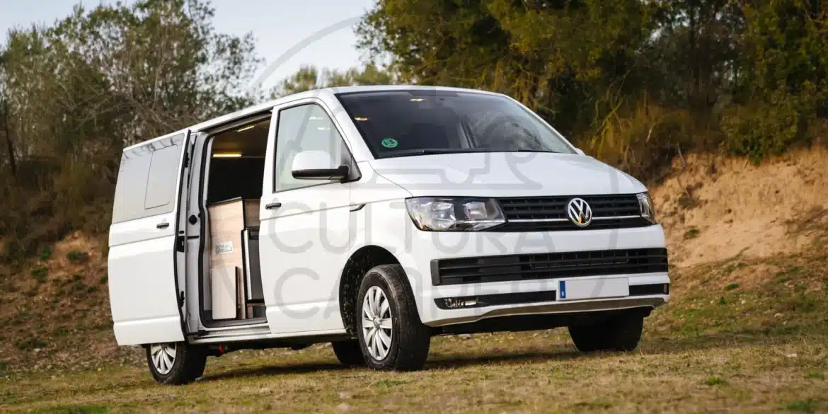 VW Caravelle T6 Camper auf einer Wiese mit geöffneter Schiebetür, Campingausbau im Innenraum sichtbar.