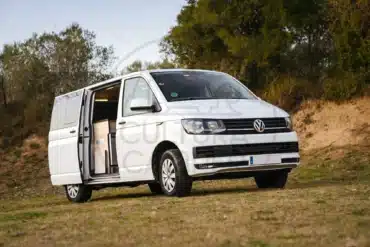 VW Caravelle T6 Camper auf einer Wiese mit geöffneter Schiebetür, Campingausbau im Innenraum sichtbar.