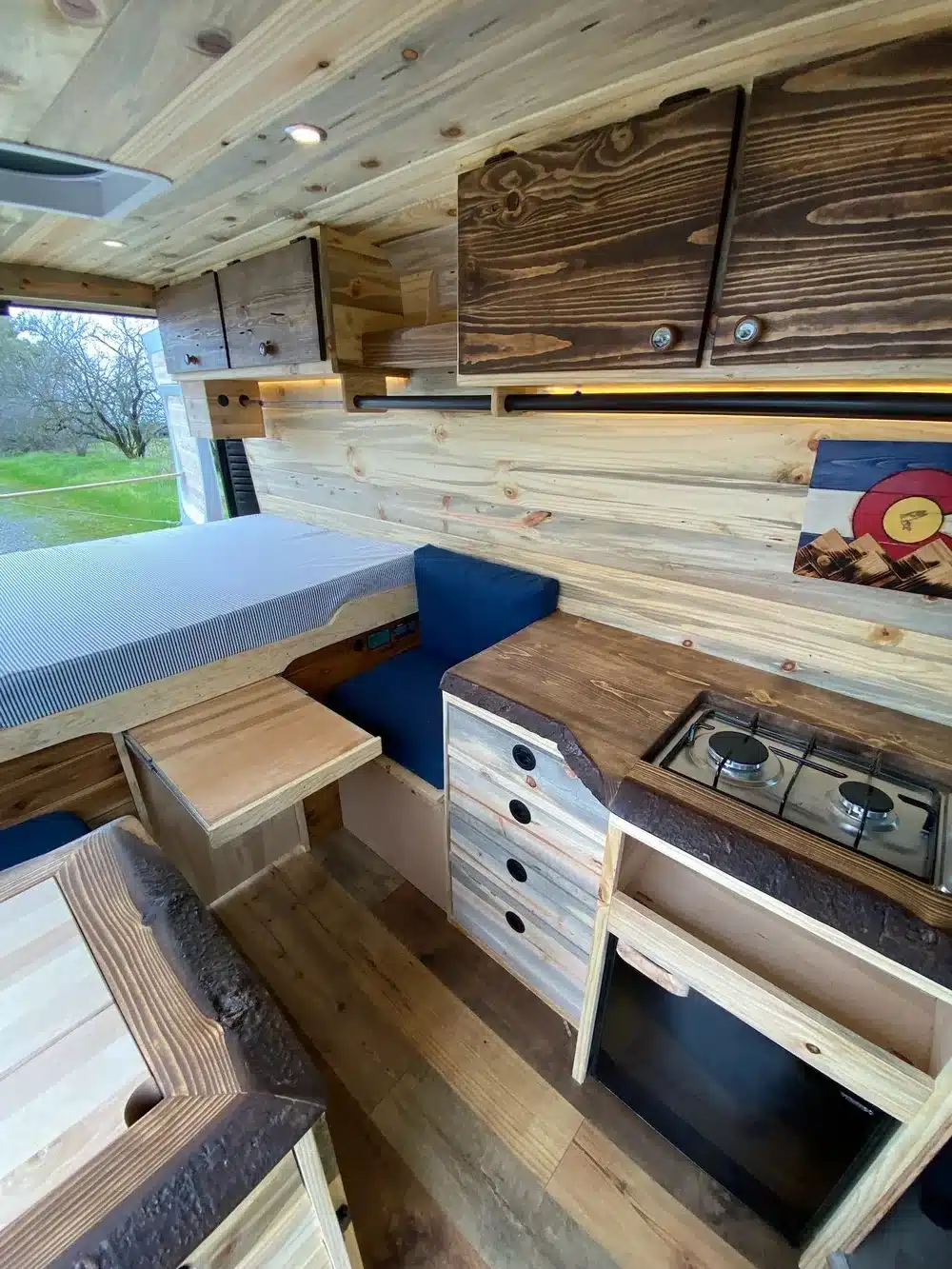 Holzverkleideter Innenraum eines Yolo Cozy Cabin Campervans mit Bett, Sitzgelegenheit, Tisch, Schränken und Gaskocher