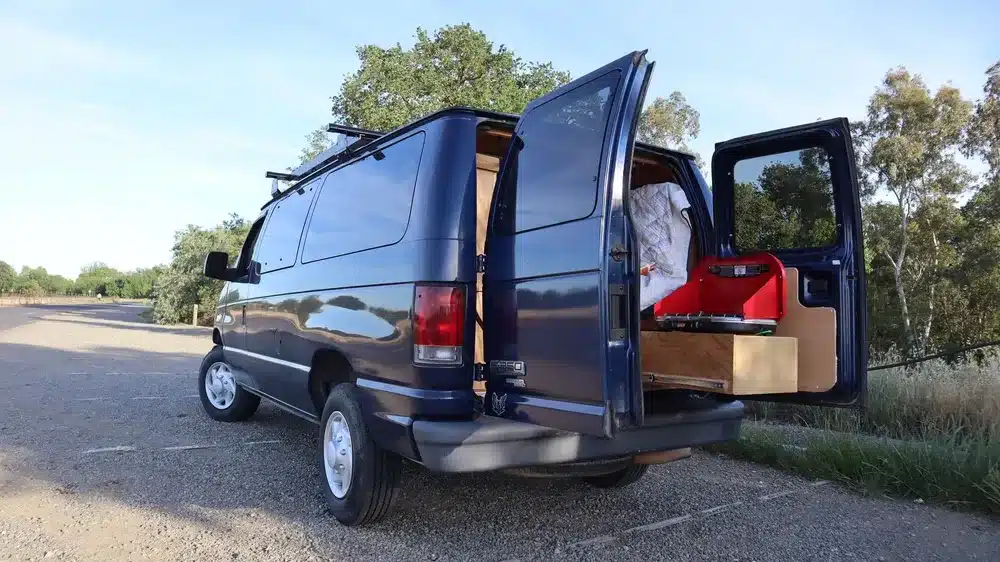 Der geöffnete Donovan Campervan auf einem Feldweg, mit zugänglichem Stauraum und Einblick in das Bett und die Outdoor-Küche.