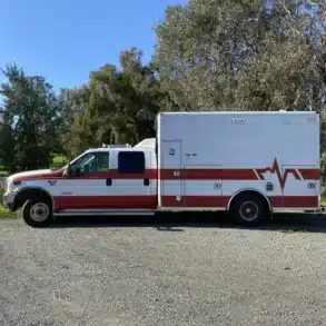 US-Ambulanzfahrzeug mit rotem Streifen, stehend auf einem Parkplatz vor Bäumen, Basis für Camper-Ausbau Fambulance.