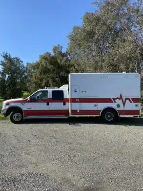 US-Ambulanzfahrzeug mit rotem Streifen, stehend auf einem Parkplatz vor Bäumen, Basis für Camper-Ausbau Fambulance.