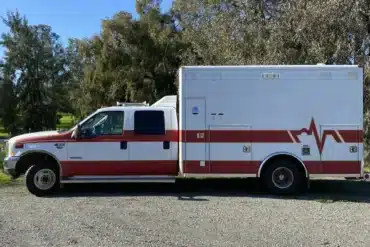 US-Ambulanzfahrzeug mit rotem Streifen, stehend auf einem Parkplatz vor Bäumen, Basis für Camper-Ausbau Fambulance.