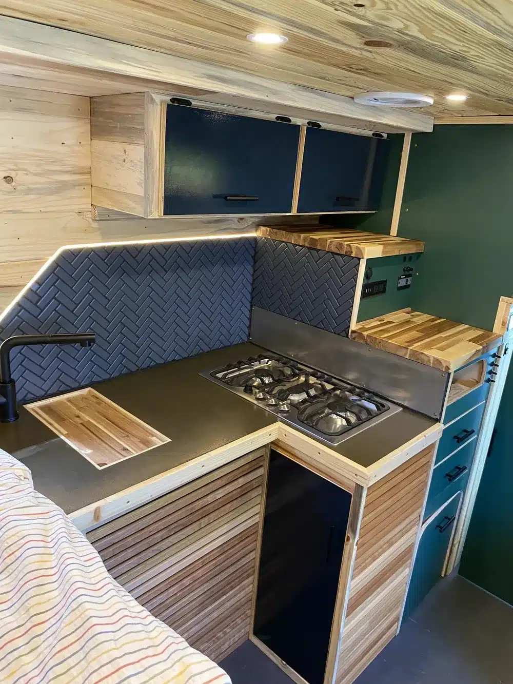 Küchenzeile im Innenraum eines als Camper ausgebauten Ambulanzfahrzeugs mit Gasherd, Spüle, Holzverkleidung und Stauraum.