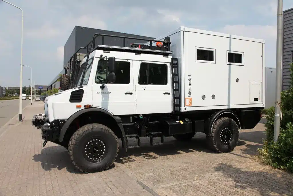 Weiße Bliss Mobil 11-Fuß-Expeditionskabine auf einem Unimog 4x4-Lkw, geparkt auf einer Straße vor Industriegebäuden.