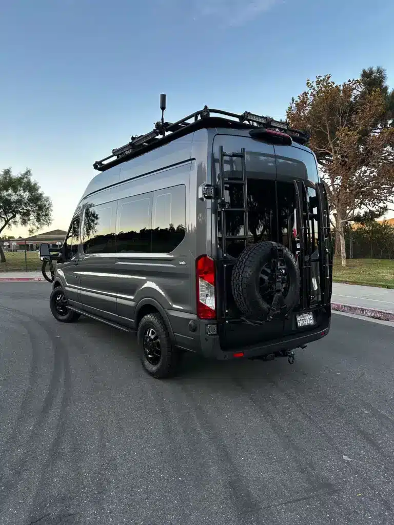 Ford Transit 350 HR PASS XL AWD Overland Van, Baujahr 2022, in dunkelgrau, mit Dachträger, Leiter und Ersatzradträger, aufgenommen auf einem Parkplatz.