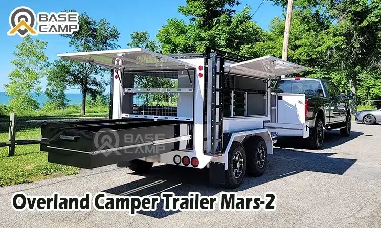 Base Camp Mars 2 Zeltanhänger angehängt an einen Pickup, mit ausgeklappten Staufächern und Küchenmodul auf einem Parkplatz im Grünen.