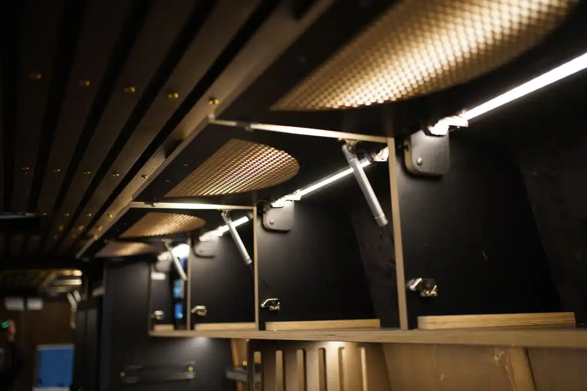 Innenaufnahme eines Campers mit offenen, beleuchteten Oberschränken und eleganter Holzverkleidung.