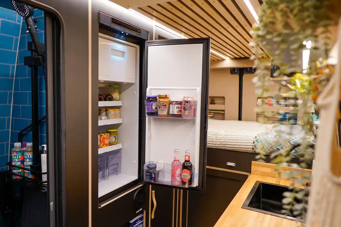 Geöffneter Kühlschrank im Beyond4 Campervan zeigt Lebensmittel und Getränke, daneben Spülbecken, Bett und moderne Innenausstattung.