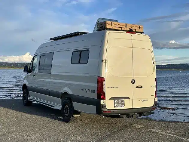 Ein heller Campingbus mit Dachbox geparkt am Wasser, im Hintergrund Landschaft und Himmel