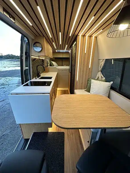 Innenansicht eines modernen Campervans mit Küchenzeile, Tisch und Sitzplätzen, Holzverkleidung an Decke und Wand.