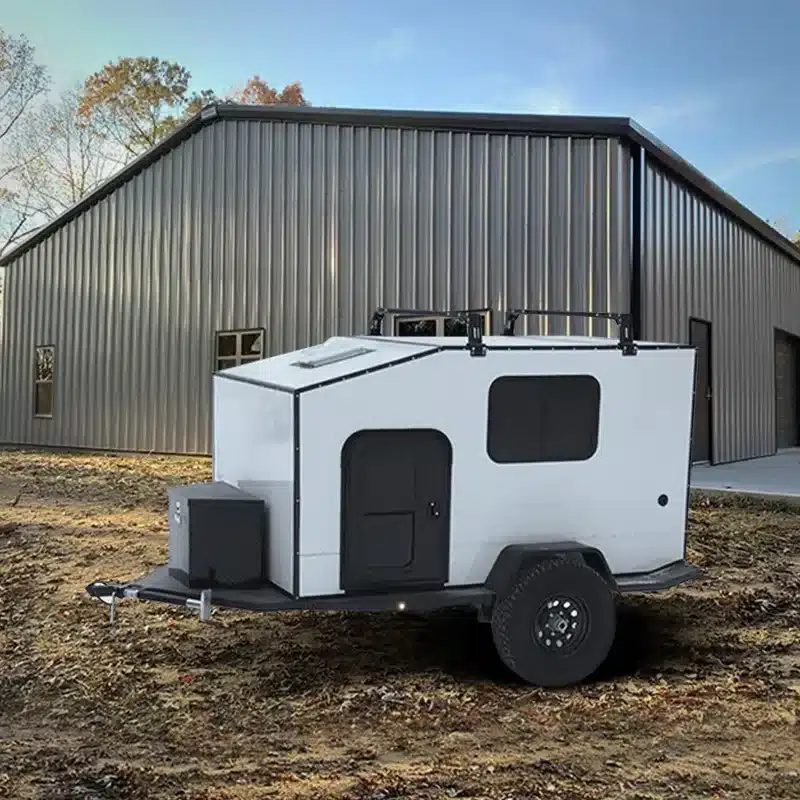Black Rhino MINI 2E Offroad Caravan auf einem Grundstück vor einer grauen Metallhalle, Seitenansicht, Offroad-Bereifung, kompakte Bauweise