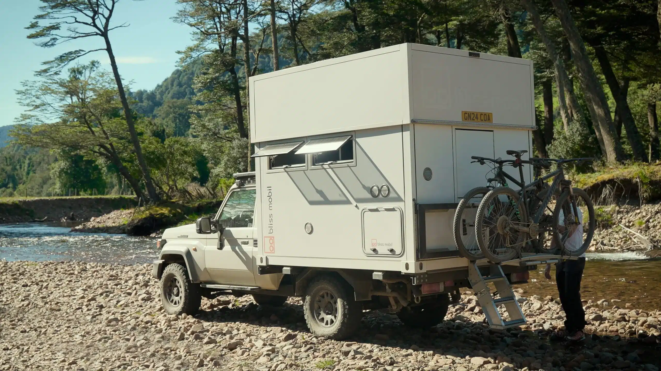 Bliss Mobil 8 Foot Expeditionskabine auf Pick-up mit ausgefahrenem Hard-Side-Hubdach und zwei Fahrrädern am Heckträger am Flussufer