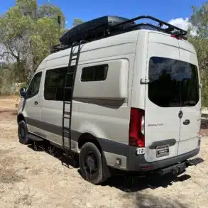 Seitenansicht eines Mercedes Sprinter 4x4 Adventure-Vans mit Dachträger, Staubox, Leiter und Seitentritt im Gelände.
