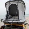 Hartschalen-Dachzelt Urban Badger montiert auf dem Dach eines gelben Fahrzeugs am Strand, geöffnet und bereit zum Camping.