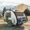 Comma CA Teardrop Camper Trailer mit geöffneter Heckklappe und Einblick in die Outdoor-Küche, Natur und Berge im Hintergrund.