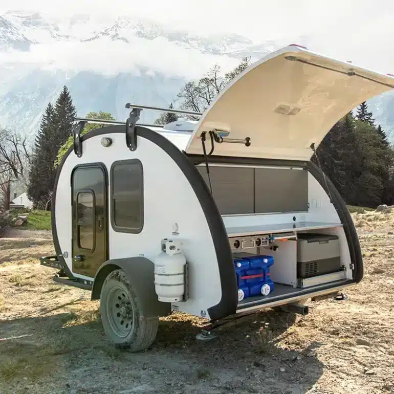 Comma CA Teardrop Camper Trailer mit geöffneter Heckklappe und Einblick in die Outdoor-Küche, Natur und Berge im Hintergrund.