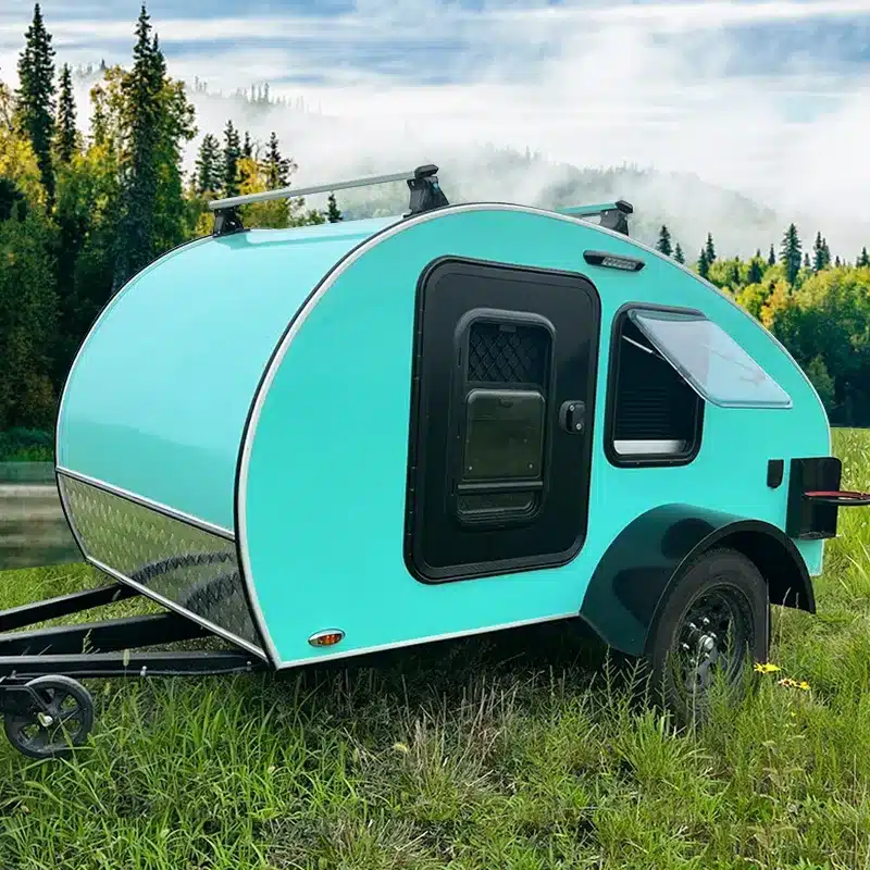 Kleiner türkisfarbener Teardrop-Anhänger Comma CC auf einer grünen Wiese vor Wald und Nebel