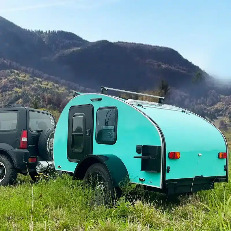 Türkiser Comma CC Teardrop-Anhänger an einem schwarzen SUV vor bewaldeten Bergen auf einer Wiese