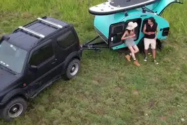 Kompakter teardropförmiger Comma CC Camping-Anhänger hinter einem SUV, Surfbrett auf dem Dach, zwei Camper genießen die Natur