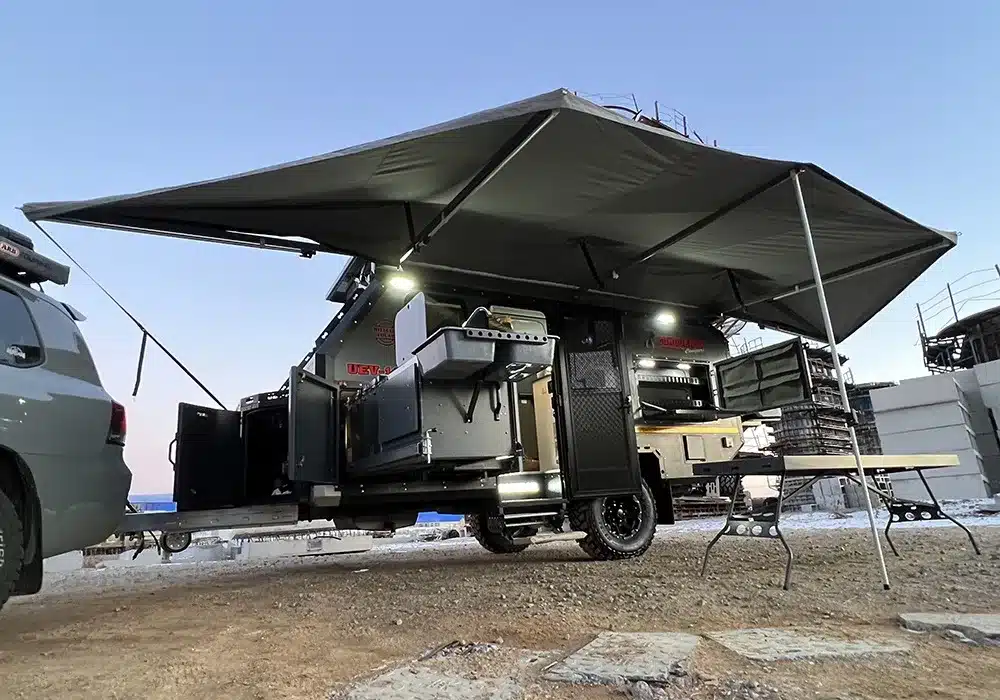 Geländegängiger Offroad-Trailer Conquer 590 mit ausgeklappter Außenküche, Markise und Stauraum, angeschlossen an ein Zugfahrzeug, auf unbefestigtem Gelände.