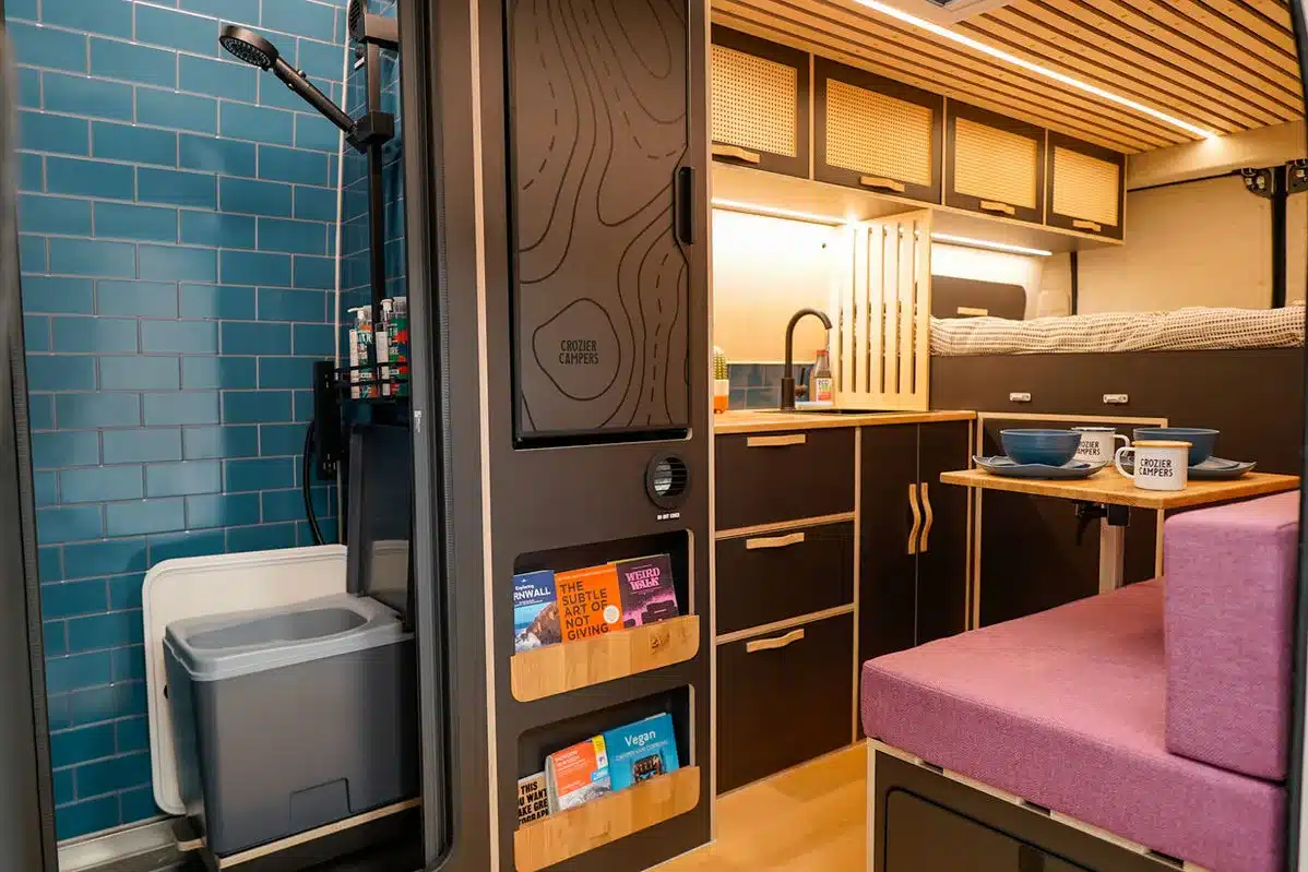 Moderner, individuell gestalteter Innenraum eines Campervans mit blauer Dusche, Küchenzeile, Sitzgruppe und Hochbett