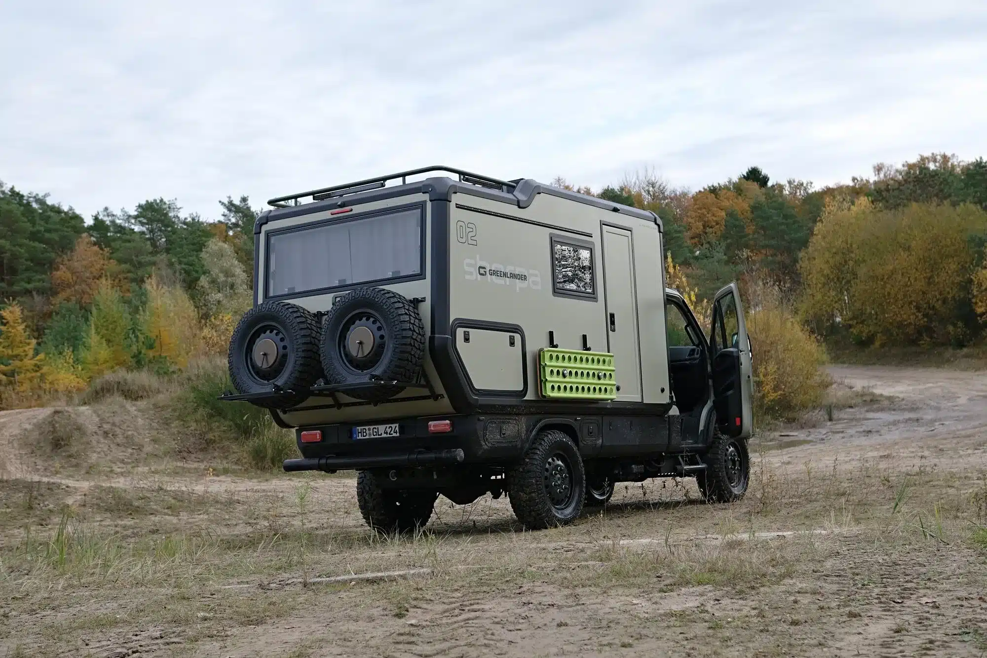 Greenlander Sherpa Expeditionsmobil mit zwei Reserverädern, auf sandigem Untergrund, umgeben von herbstlichem Wald.