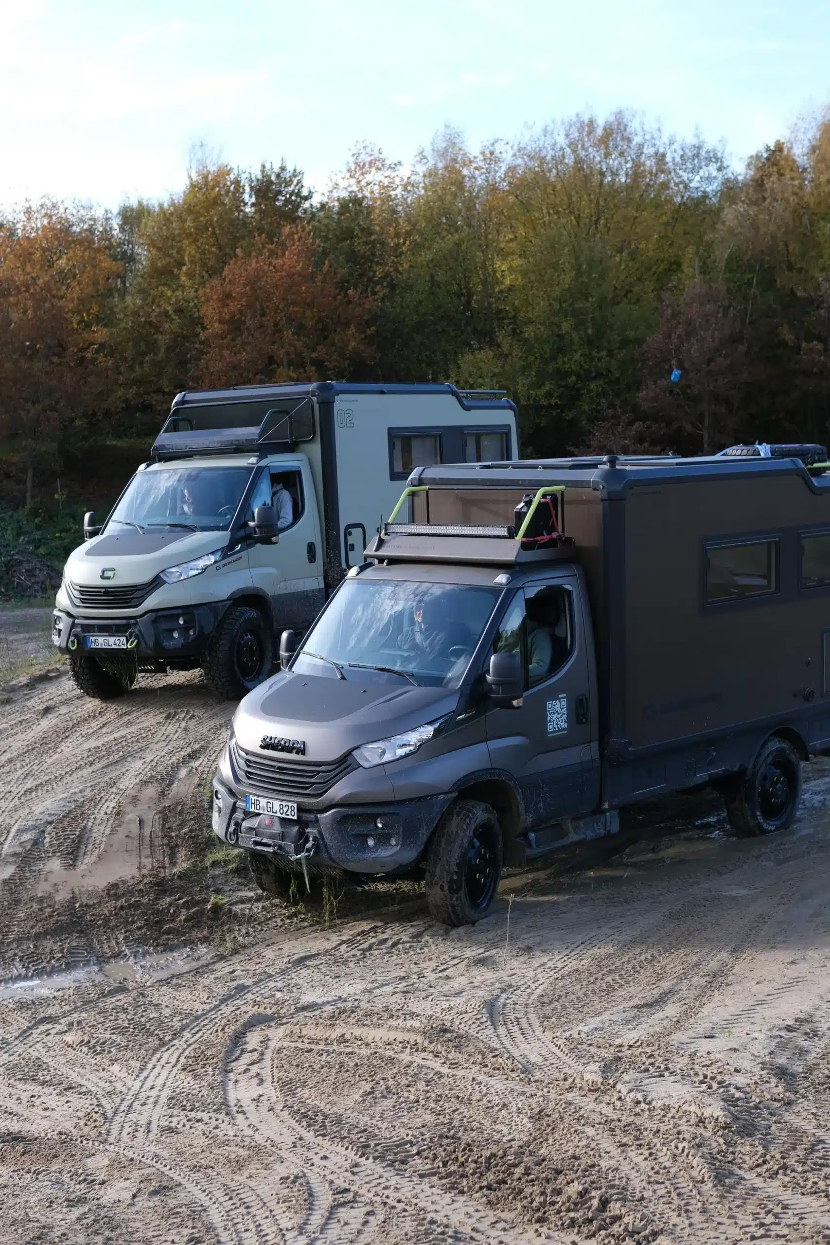 Zwei Greenlander Sherpa Expeditionsmobile fahren auf matschigem Gelände vor herbstlichem Wald.