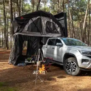 Hotomobil Atlas Cabin Box auf einem VW Pickup, ausgeklappt als Campinglösung mit Zelt und Feuerstelle im Wald.