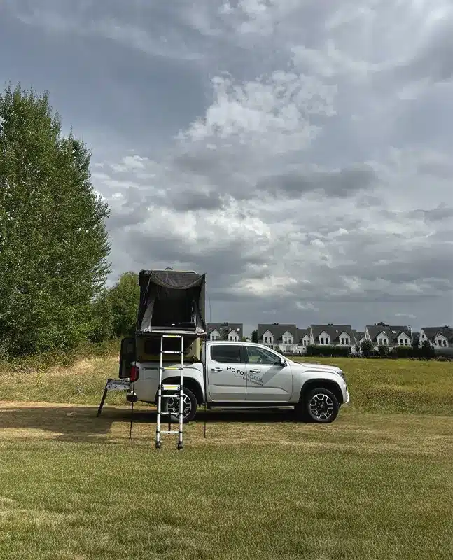 Pickup mit montierter Hotomobil Atlas Cabin Box und aufgebautem Dachzelt auf einer Wiese, im Hintergrund Wohnhäuser und Bäume.
