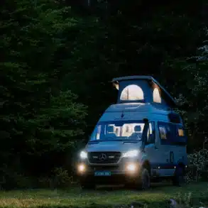 IPAR NORANTZ Campervan auf Mercedes-Basis mit aufgeklapptem Dach steht im Grünen, abends beleuchtet.