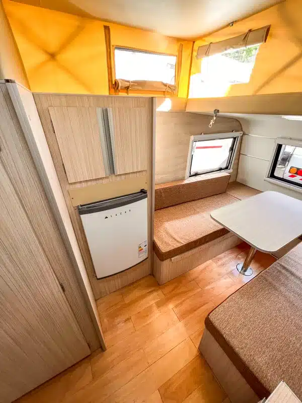 Innenraum des Itati 380 TLi mit Sitzgruppe, Tisch, Schränken und 74-Liter-Kühlschrank unter dem Hubdach.