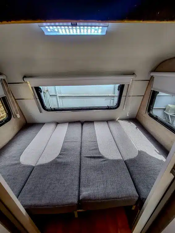 Innenansicht des Itati 380 TLi Wohnwagens: umgebautes Doppelbett mit grauen Polstern, große Fenster und LED-Licht.
