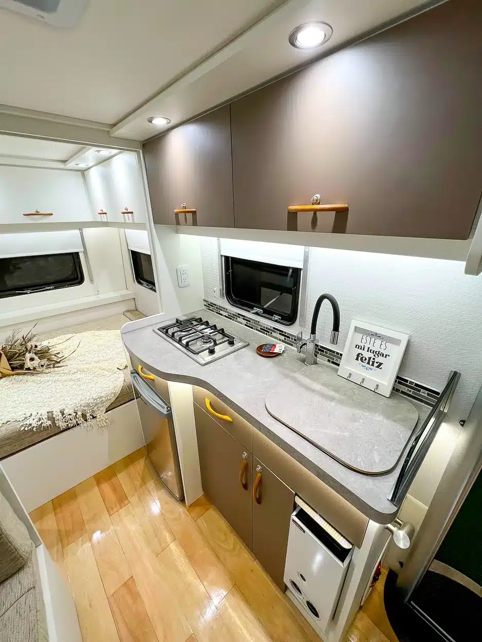 Küche und Schlafbereich im ITATI Avant Garde Pampa Wohnwagen mit Gaskocher, Spülbecken, Kühlschrank und Einblick ins Doppelbett.