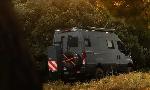 Heckansicht eines grauen Iveco Daily 4x4 L3H2 Campers mit Reserverad, abseits der Straße geparkt vor Bäumen.