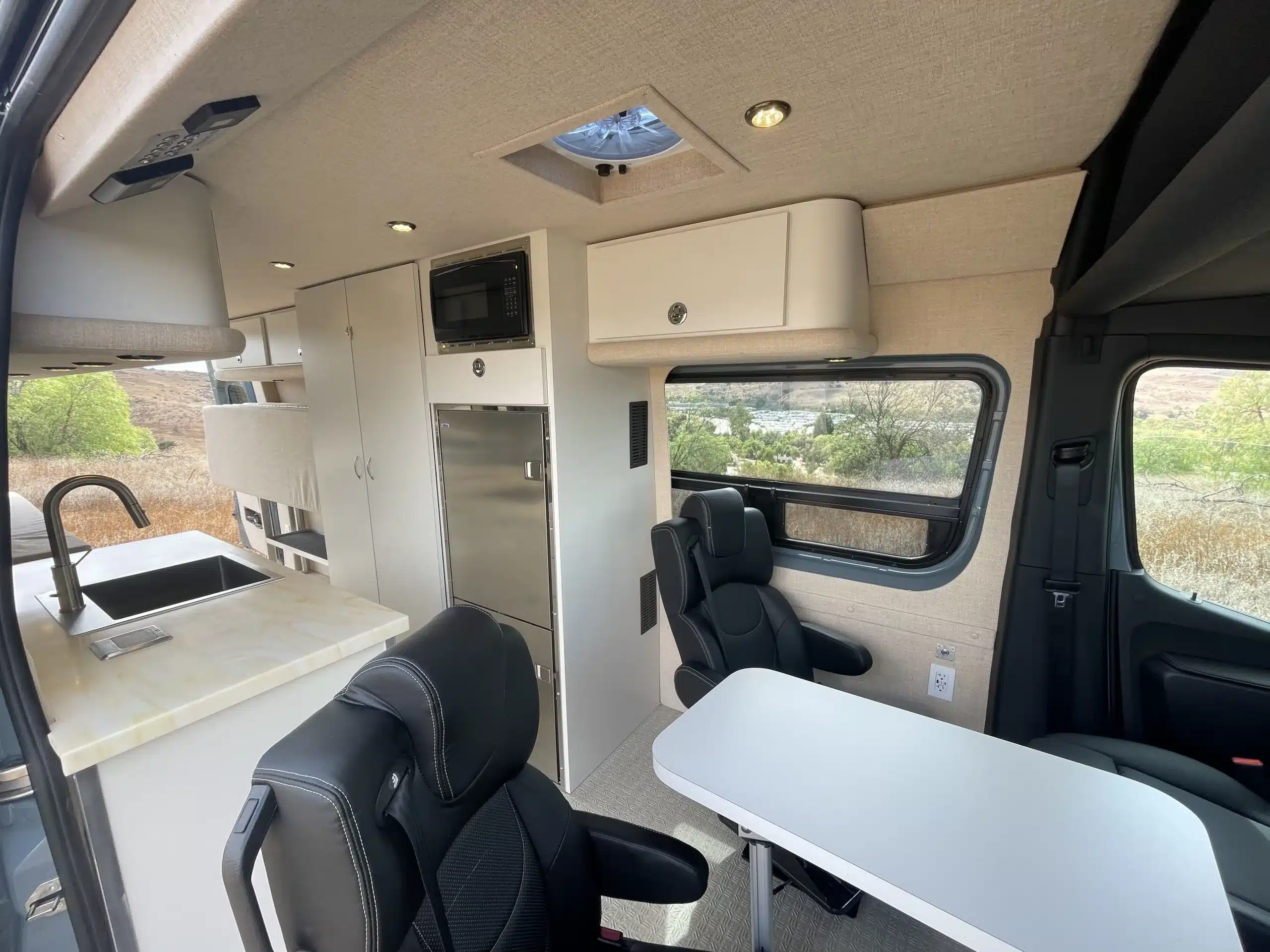 Blick in den Innenraum eines ausgebauten Jackson 4x4 Sprinter Vans mit Küchenzeile, Spüle, Mikrowelle, Kühlschrank und zwei drehbaren Captain’s Chairs an einem Tisch.