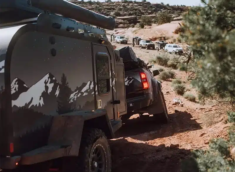 Offroad-Campertrailer, gezogen von einem Pickup, fährt auf einer matschigen, unbefestigten Strecke mit weiteren Fahrzeugen im Hintergrund