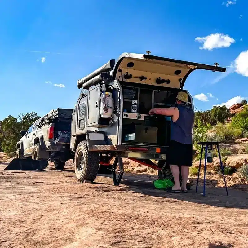JR Black Rhino VIC Offroad-Camper Trailer mit geöffneter Heckküche und Nutzer beim Outdoor-Kochen auf felsigem Untergrund.