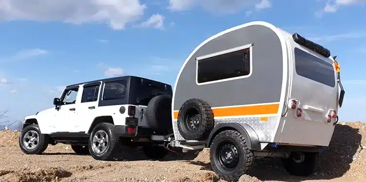 Ein weiß-grauer Kalala Teardrop Camper Trailer mit orangefarbenem Streifen wird von einem weißen Geländewagen im Gelände gezogen
